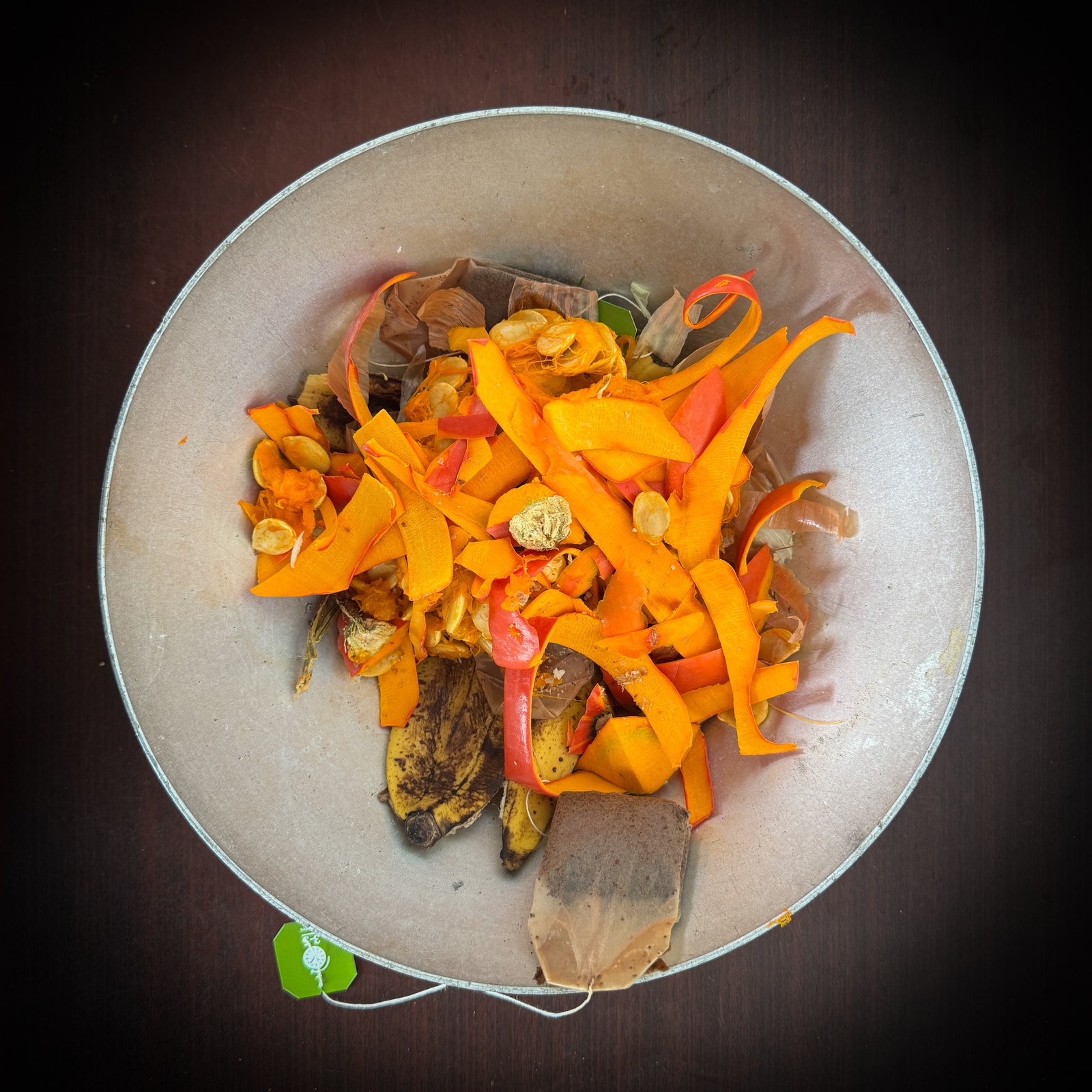 A metal bowl filled with chopped orange and red vegetables, banana peels, and other kitchen scraps sits on a dark wooden surface.