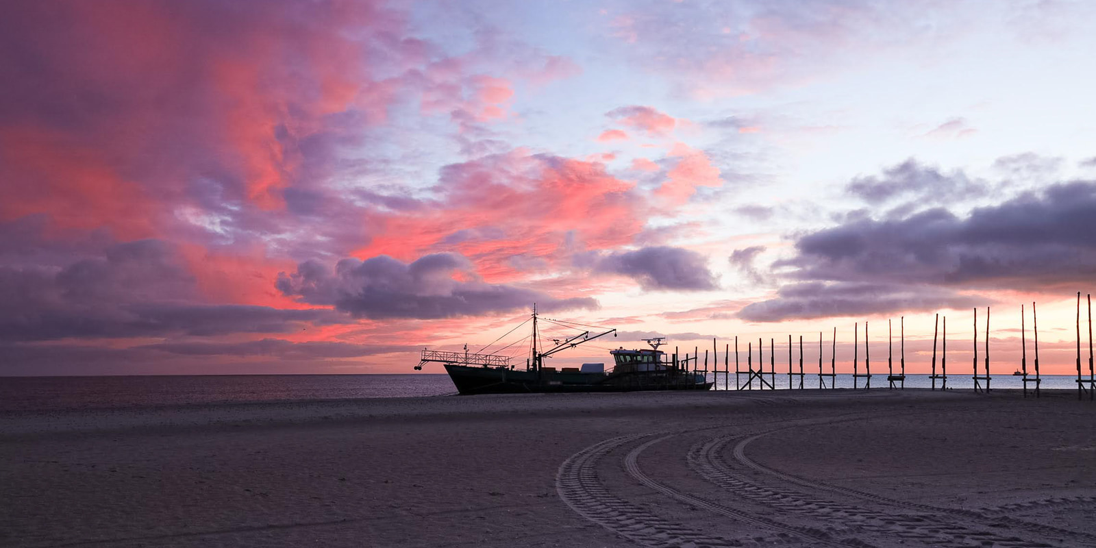 A fishing boat rests on a sandy beach at sunset, with a dramatic sky of pink and purple clouds above.