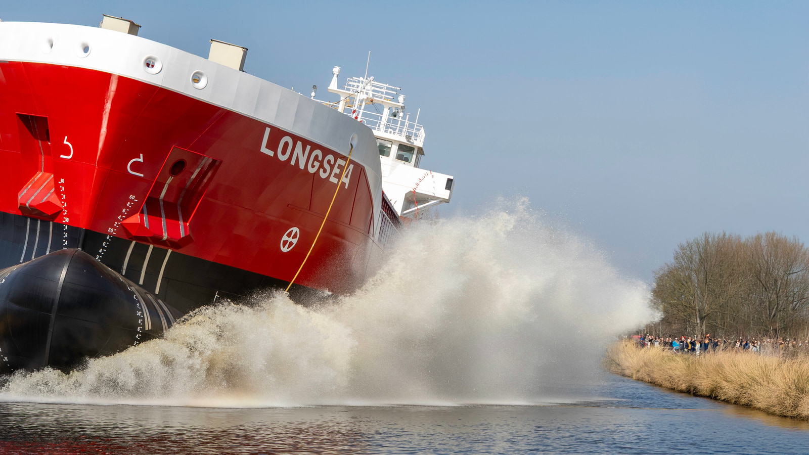 A large red and white cargo ship labeled "LONGSEA" pushes through a body of water, creating a dramatic spray of water as it moves forward.