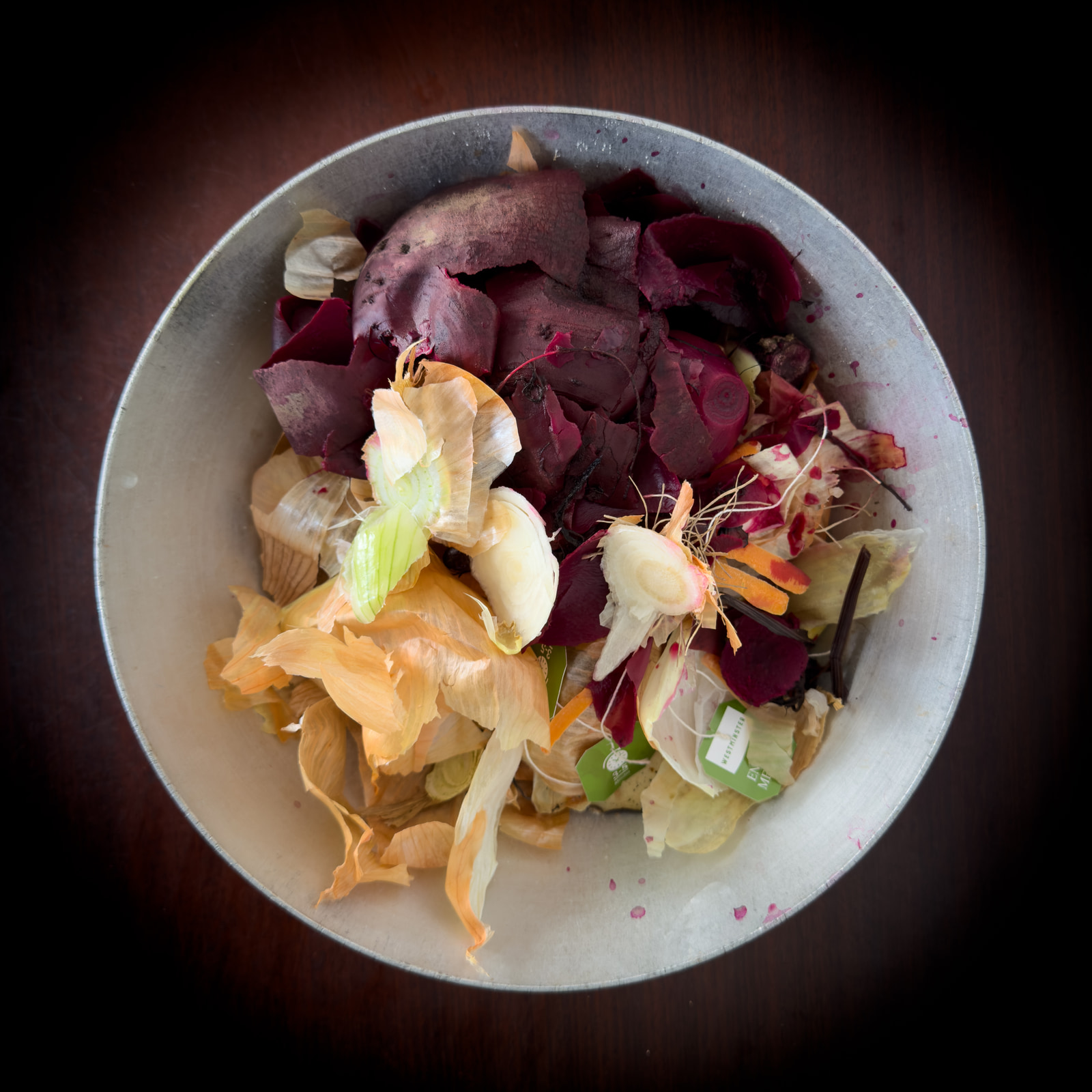 A metal bowl filled with a colorful assortment of vegetable scraps, including beet leaves, onion skins, and carrot peels, arranged on a dark wooden surface.