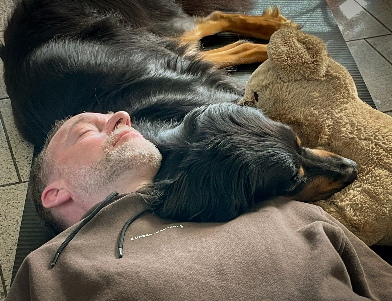 A man rests his head on a soft surface with two dogs lying beside him, all appearing relaxed on a tiled floor.