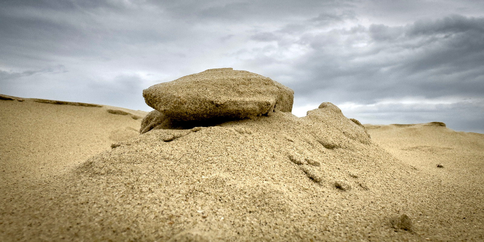 A large, weathered sandstone boulder rests atop a dune in a desert landscape under a cloudy sky.