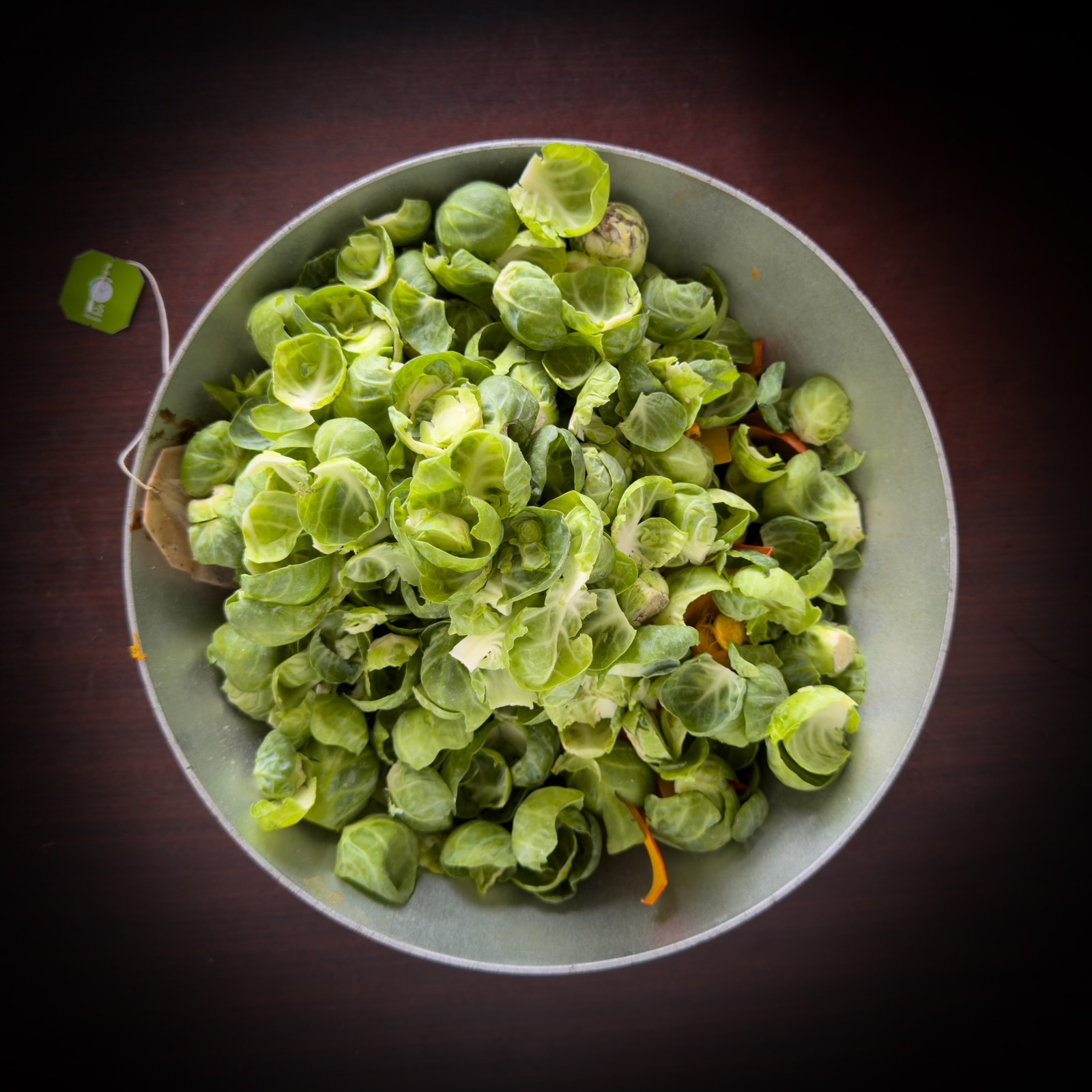 A metal bowl filled with raw Brussels sprouts sits on a dark wooden surface.