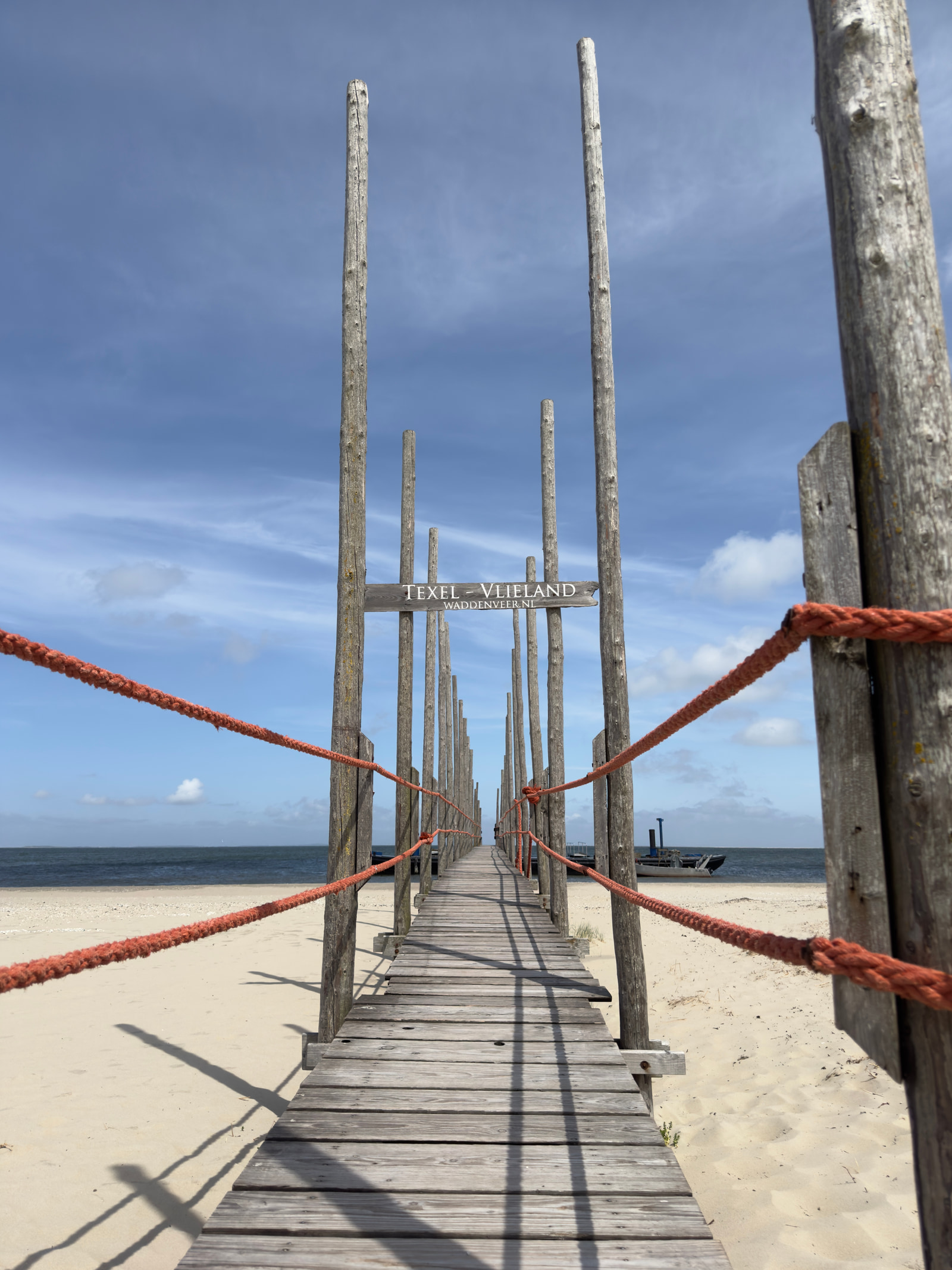 A wooden boardwalk extends over sandy beach toward the ocean, bordered by red ropes and wooden posts.